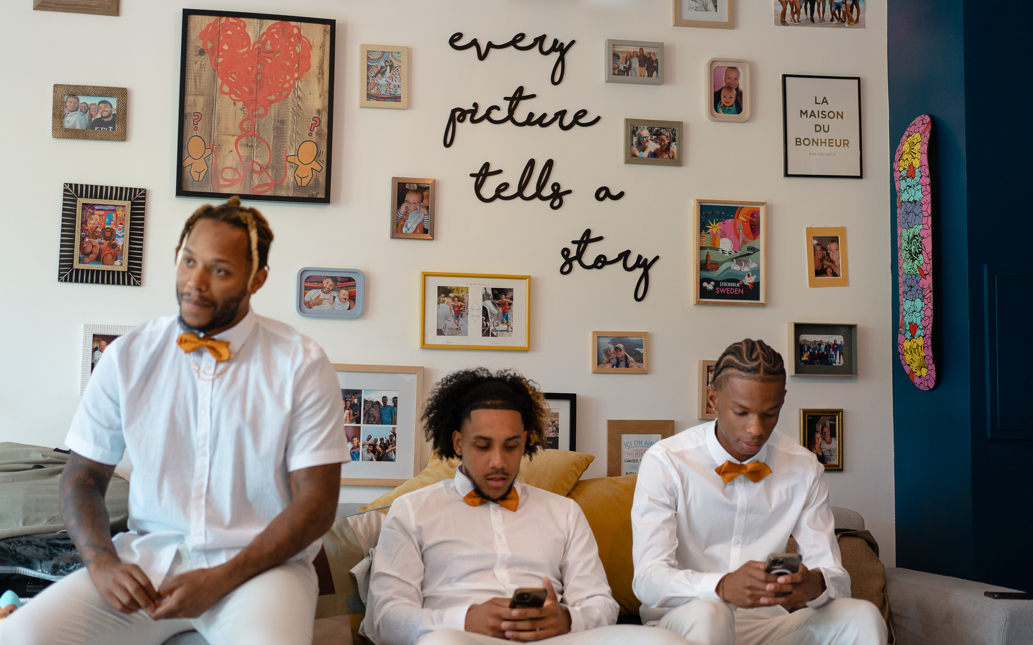 Trois amis du marié sont assis sur le canapé. L'un est flou et les deux autres, net, jouent sur leurs téléphones. L'arrière plan est le mur photo de la maison où le marié se prépare et il est écrit "every picture tells a story"