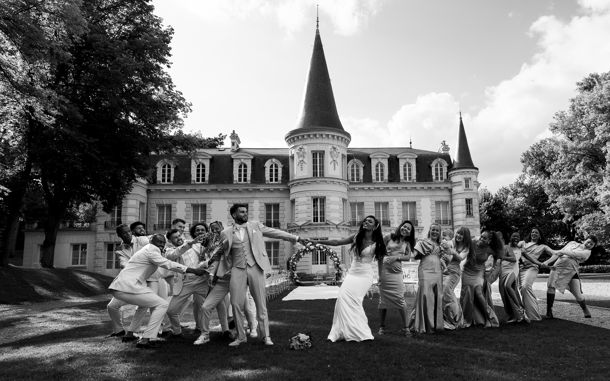 Photo de groupe devant le château du lieu de réception. Les mariés sont au centre et les témoins de chacun tirent le bras des mariés qui se tiennent la main tant bien que mal.